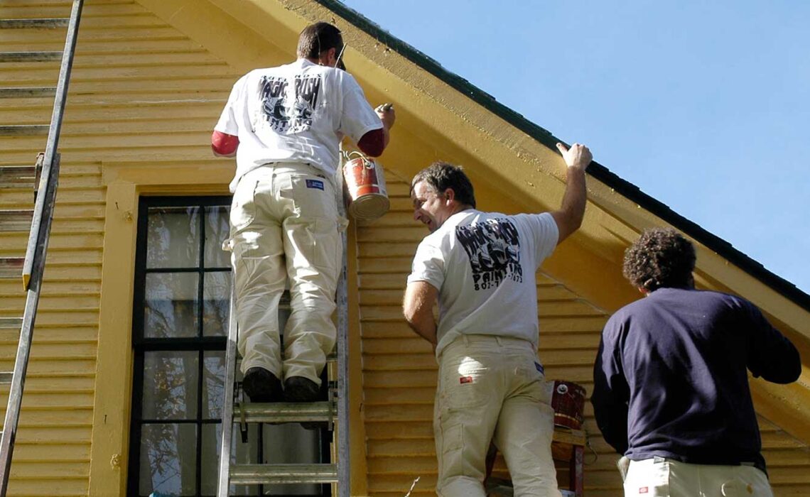 A three-man paint crew topcoats a wood-sided home exterior